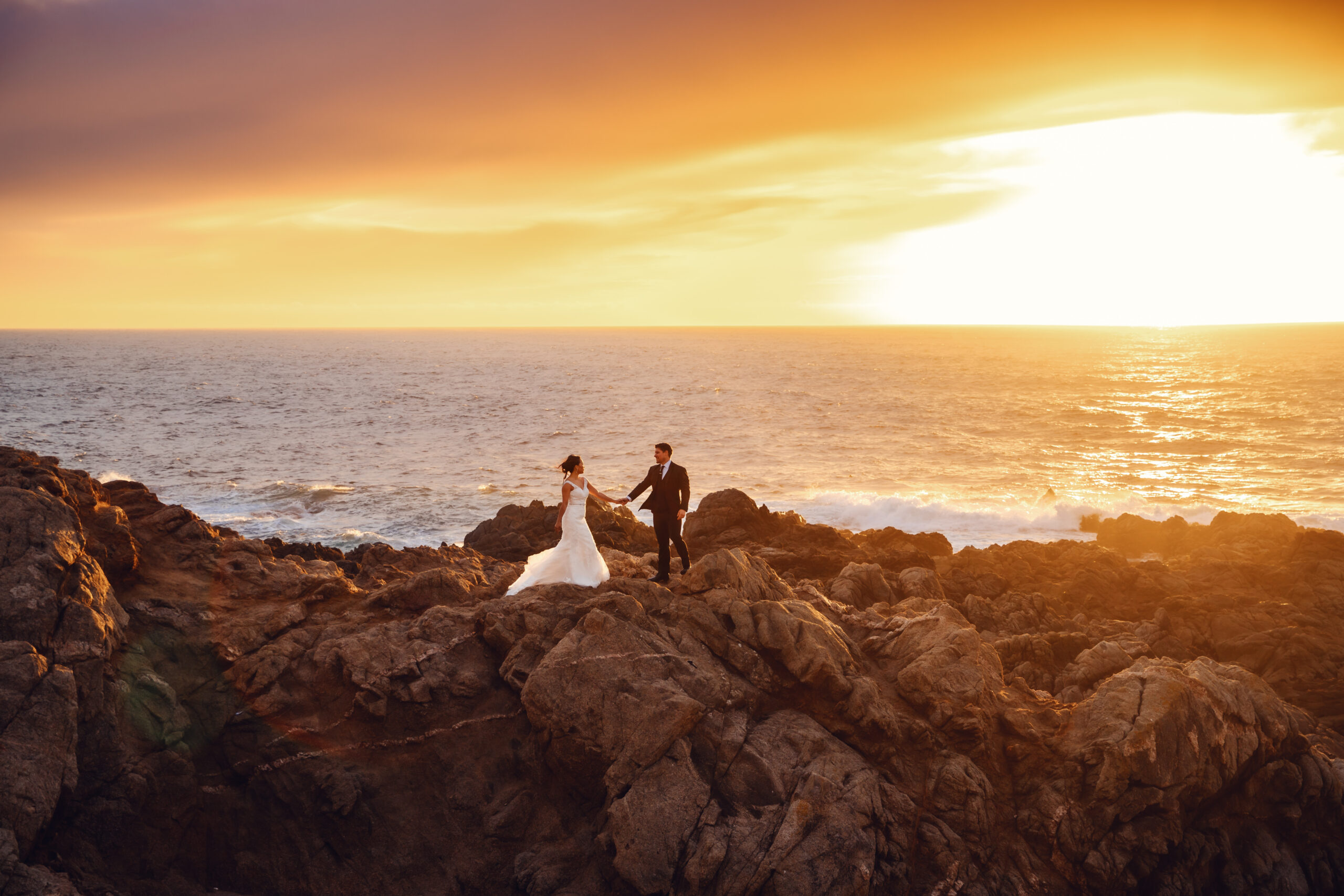 a couple holds hands on the rocky coast of Big Sur with a beautiful sunset over the ocean behind them during their Big Sur Elopement.