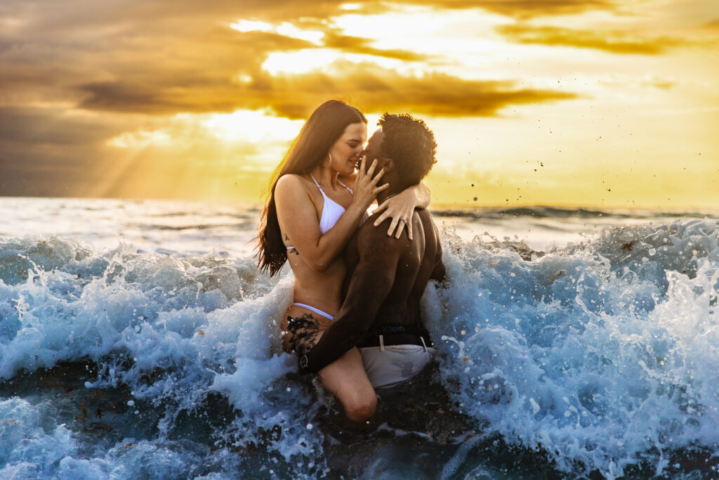 a couple kisses in the ocean as a wave crashes into them and the sun rises behind them. engagement session guide