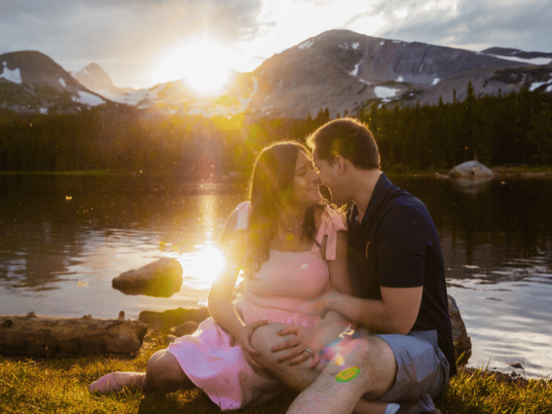 a couple cuddles in front of a lake as the sun sets behind the mountains behind them