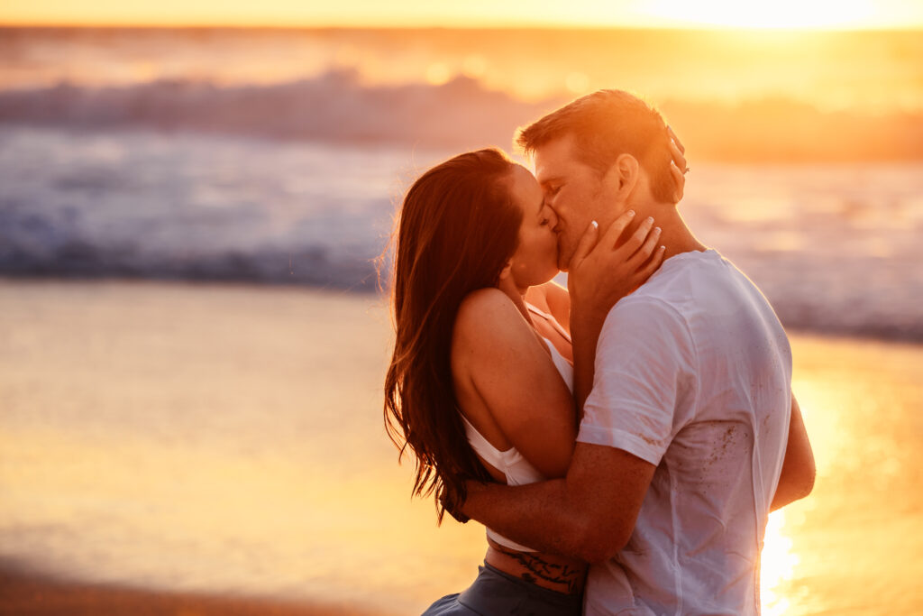 A couple eloped in Florida and shares a passionate kiss on the beach, bathed in the warm glow of the sunset as the ocean waves gently crash behind them.