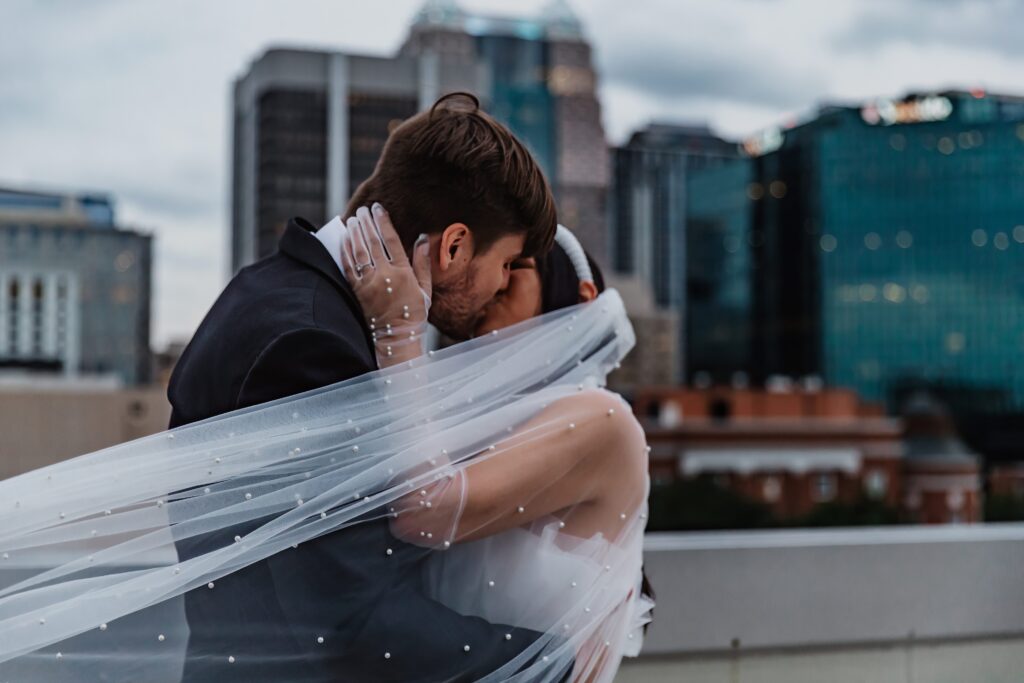 A couple eloped in Florida and shares a passionate kiss on a rooftop, with the bride’s veil flowing in the breeze and the city skyline as their backdrop.