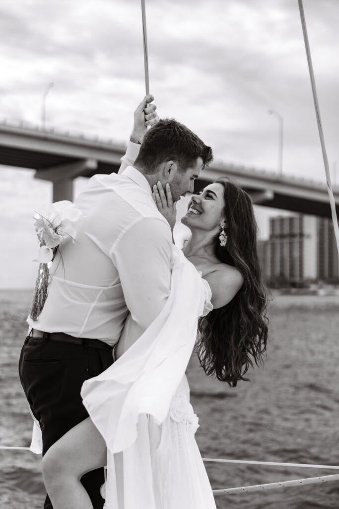 A couple eloped in Florida and shares a playful, romantic moment on a boat, laughing and kissing beneath a bridge, with the bride's flowing dress gently swaying in the breeze.