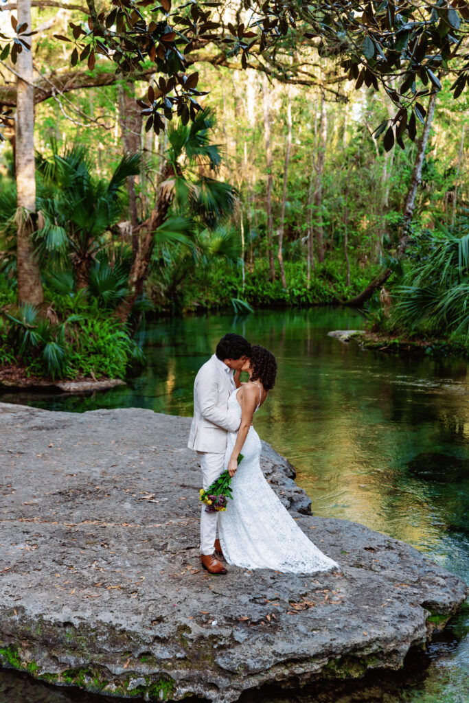 A couple shares a quiet Florida elopement moment on a limestone ledge at Ichetucknee Springs State Park, leaning in for a kiss beside the clear spring water, captured by Florida Elopement Photographer Shannon Lee.