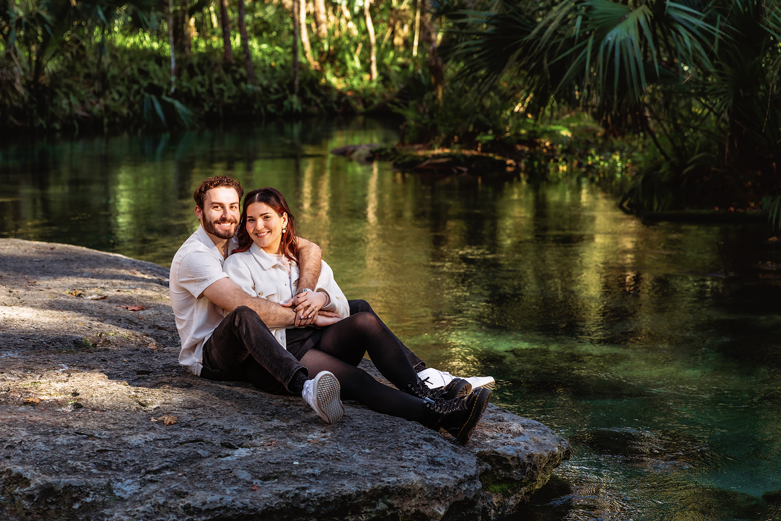 A couple sits snuggled together on a mossy limestone rock beside the crystal-clear turquoise waters of Kelly Rock Springs, surrounded by lush Florida palms — a serene and romantic Orlando elopement setting.