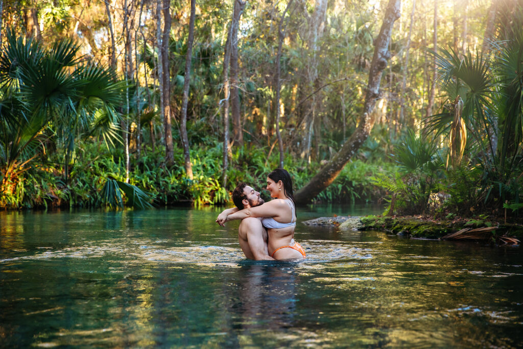 An engaged couple embraces while swimming in a clear spring surrounded by lush greenery, photographed by Orlando Engagement Photographer Shannon Lee.