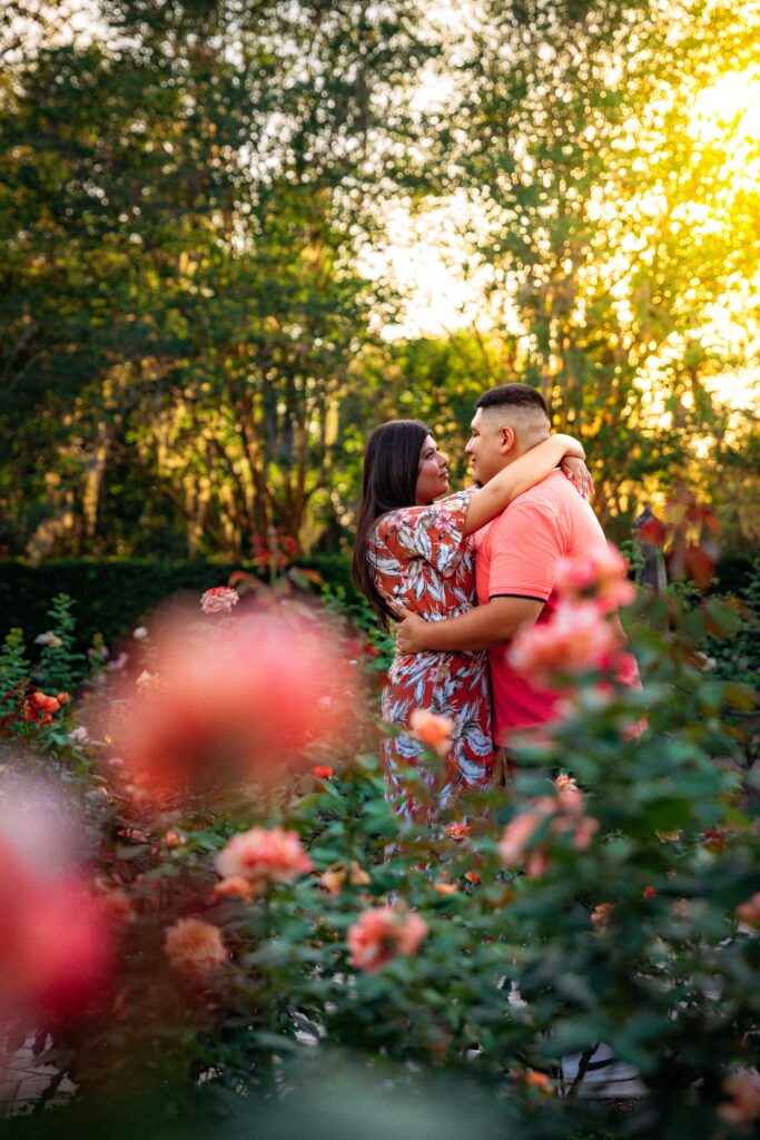 An engaged couple shares a quiet engagement moment among blooming roses at Harry P. Leu Gardens, holding each other close and gazing softly, captured by Orlando Engagement Photographer Shannon Lee.