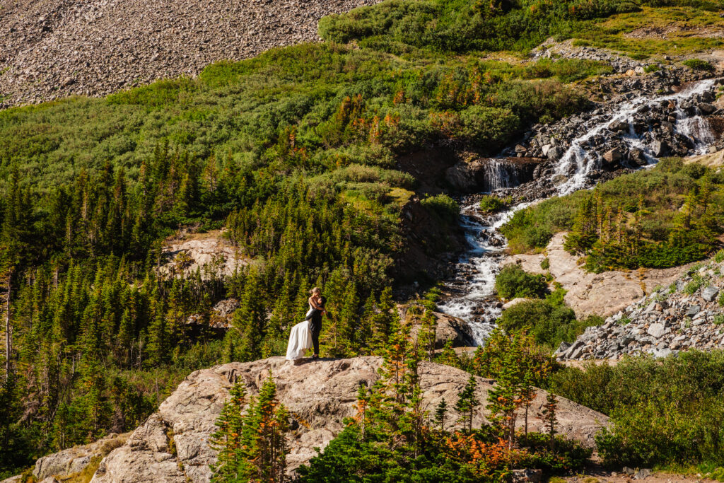 A couple embraces on a rocky ledge beside a cascading waterfall for their Breckenridge elopement in Colorado.