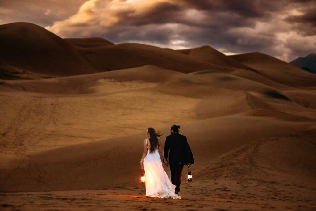 A couple walks hand in hand carrying lanterns across the dunes at dusk for their Great Sand Dunes National Park elopement in Colorado.