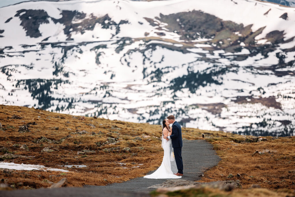 A couple kisses on a winding path with snow-capped peaks behind them for their Rocky Mountain National Park elopement in Colorado.