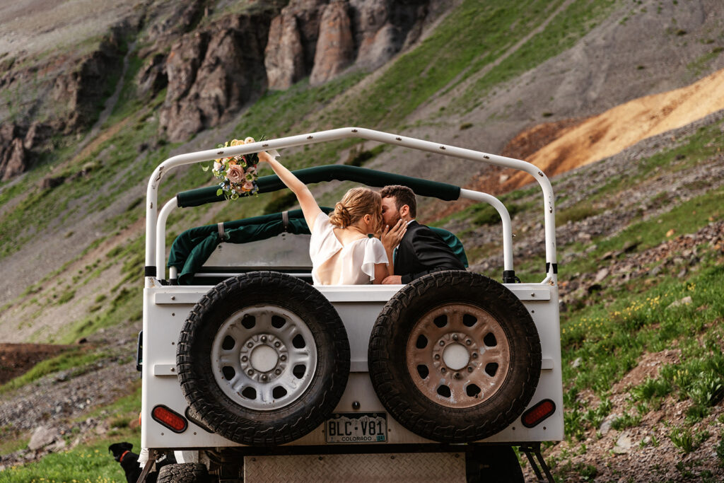 A couple has an elopement session in Colorado and shares a kiss in the back of a vintage off-road vehicle surrounded by rugged mountain scenery, photographed by Shannon Lee, their destination elopement photographer.