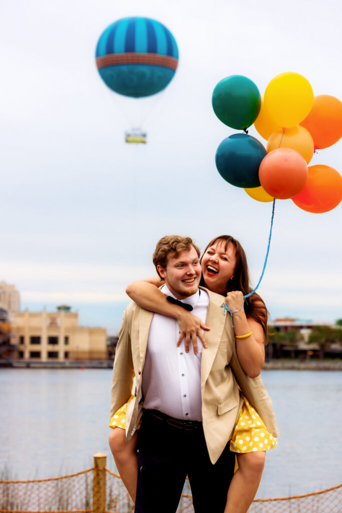 A laughing couple embraces waterside holding a cluster of colorful balloons, with the iconic Disney Springs hot air balloon floating in the background — a joyful and playful Universal Studios Orlando elopement moment.