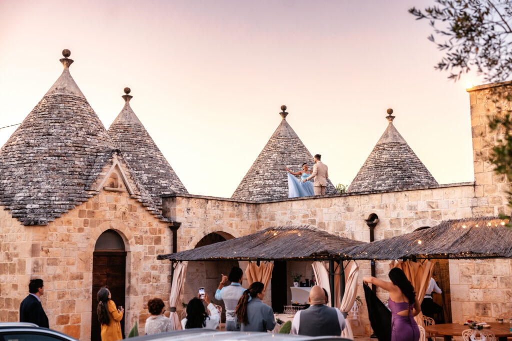 A bride in a flowing blue gown dances with her groom atop a historic trulli rooftop at golden hour, as delighted guests below look up and capture the magical moment at their Italy destination wedding in Puglia.