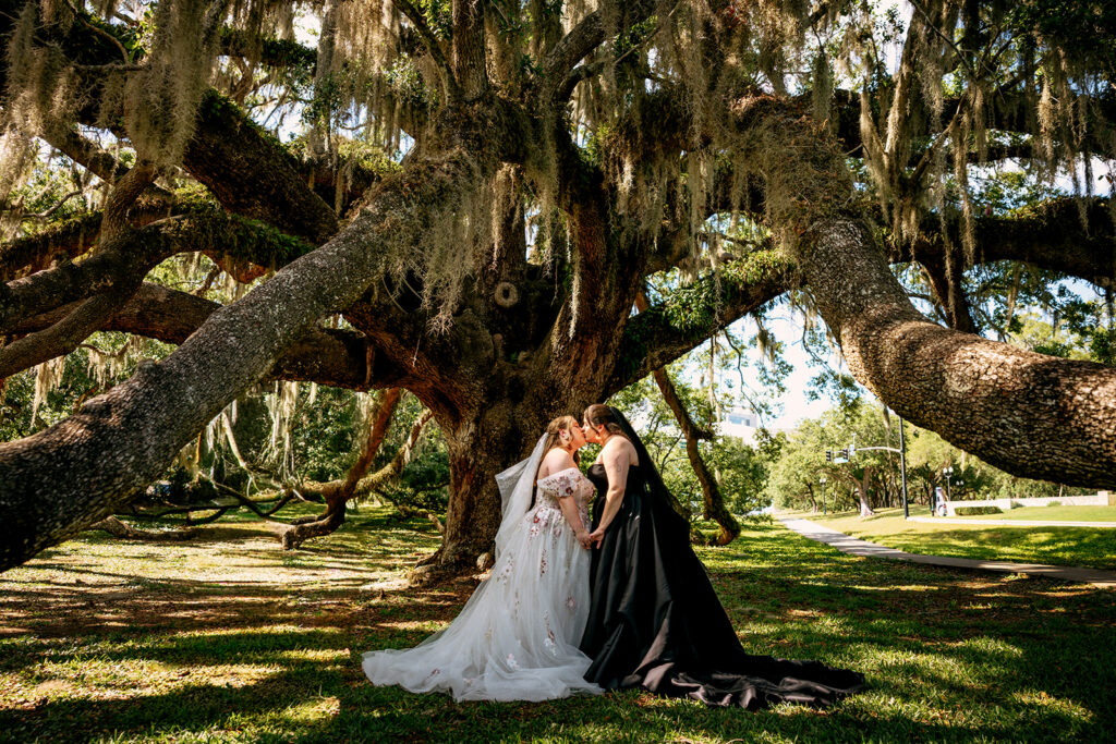 Two brides share a kiss beneath a sprawling moss-draped live oak tree — one in a floral white ball gown with veil, the other in a dramatic black gown — in a breathtaking outdoor portrait from their Orlando Science Center destination wedding.
