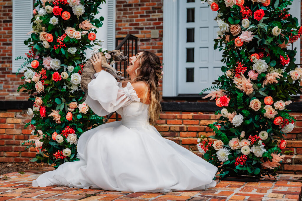 A laughing bride in a off-shoulder white gown sits on a brick courtyard floor, joyfully nuzzling her French Bulldog between two lush floral arches bursting with coral, peach, and white blooms — a perfect wedding day moment with her beloved pet.