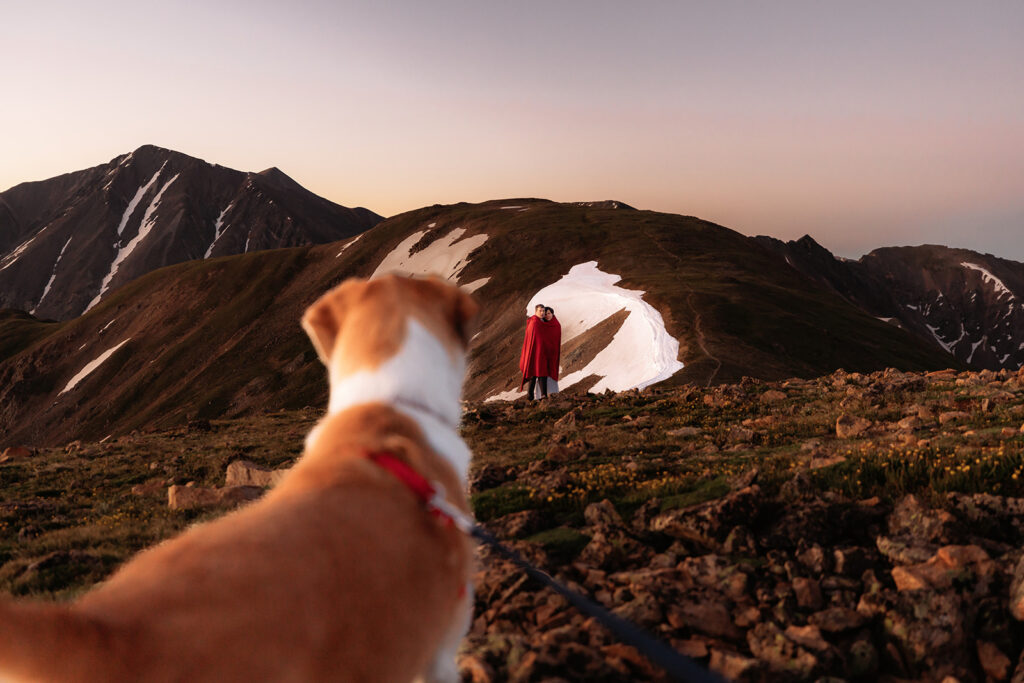 A couple wrapped in a red blanket stands together before a stunning snow-patched alpine ridge at sunset, framed beautifully through the blurred foreground of their dog on leash — a heartfelt Colorado mountain elopement with pets.