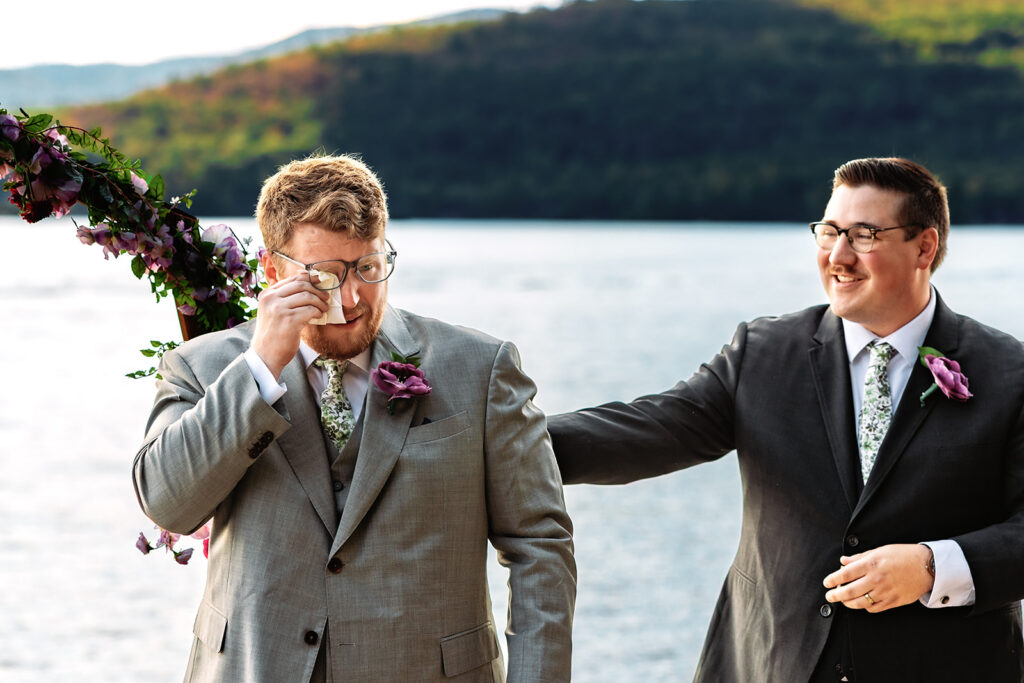 A tearful groom wipes his eyes as his smiling best man places a supportive hand on his shoulder while awaiting his bride at their lakeside Adirondack Camp destination wedding, with misty mountain views in the background.