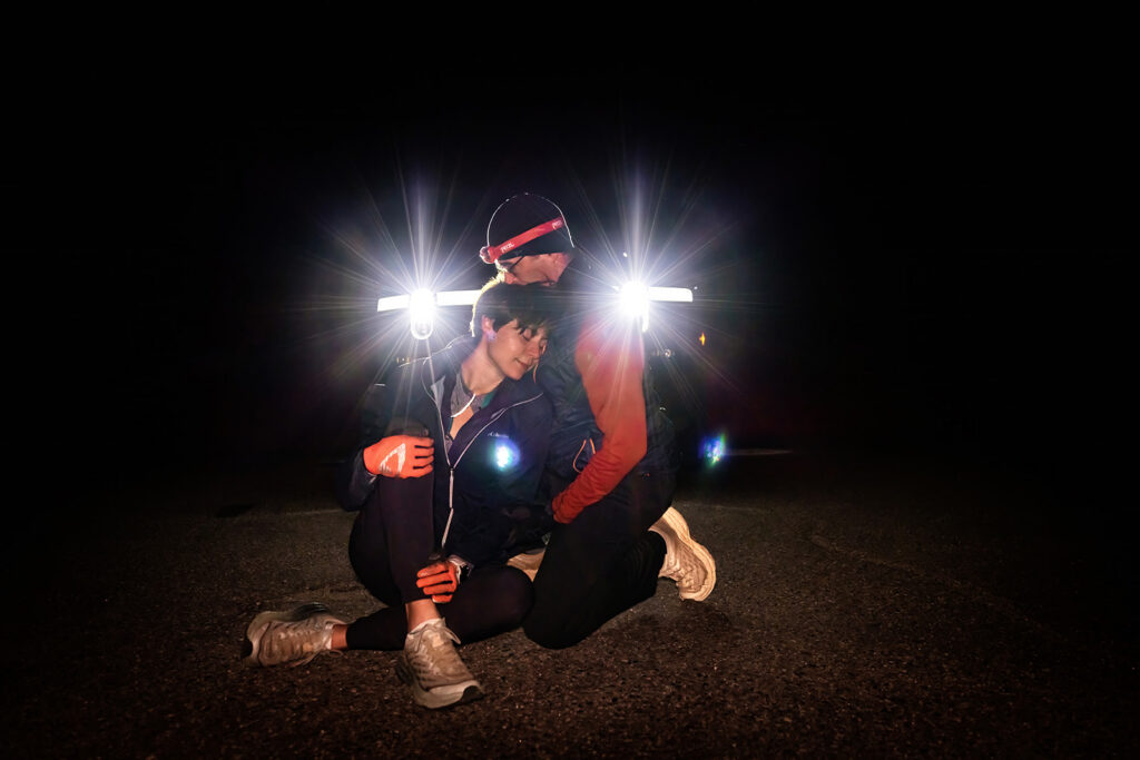 A couple sits together in the dark alpine night, wearing headlamps and hiking gear that illuminate them with dramatic star-burst light flares — a uniquely adventurous and intimate Loveland Pass elopement moment.