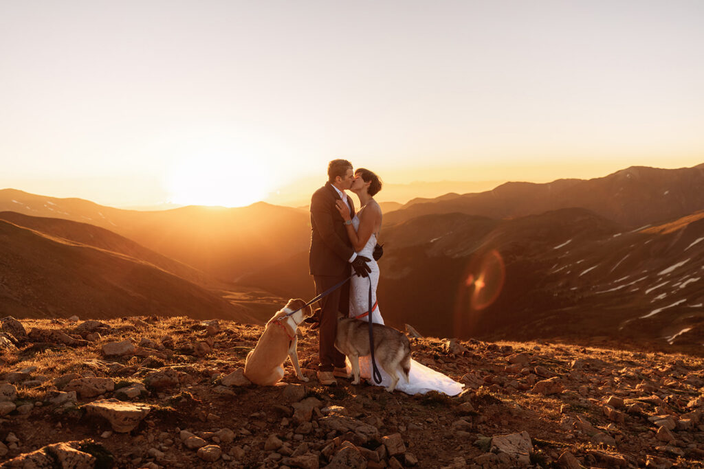 A bride and groom share a golden kiss on a rocky mountain summit at fiery sunset, their two dogs sitting loyally at their feet, in an epic and heartfelt Loveland Pass elopement with pets.