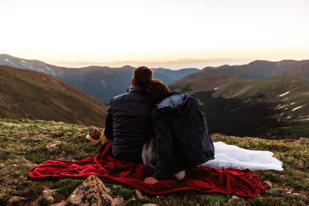 A bride and groom sit wrapped together on a red blanket with their dog, gazing in peaceful silence over a vast Colorado mountain panorama at sunset during their intimate Loveland Pass elopement.