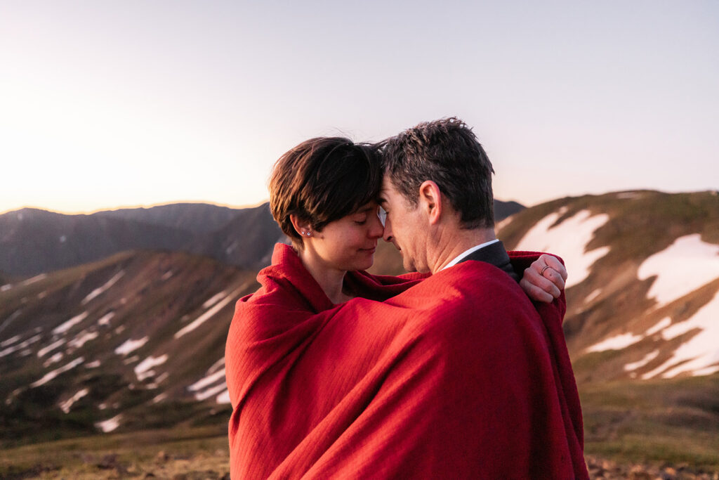 A couple wrapped together in a red blanket touch foreheads in a tender, intimate moment against a snow-streaked alpine landscape at their dreamy Loveland Pass elopement in Colorado.