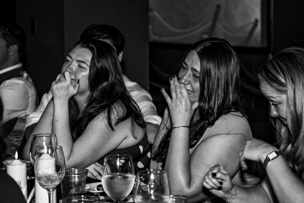 Black and white photo of women laughing and reacting with hands near their faces while seated at a candlelit table with wine glasses, during a wedding reception.