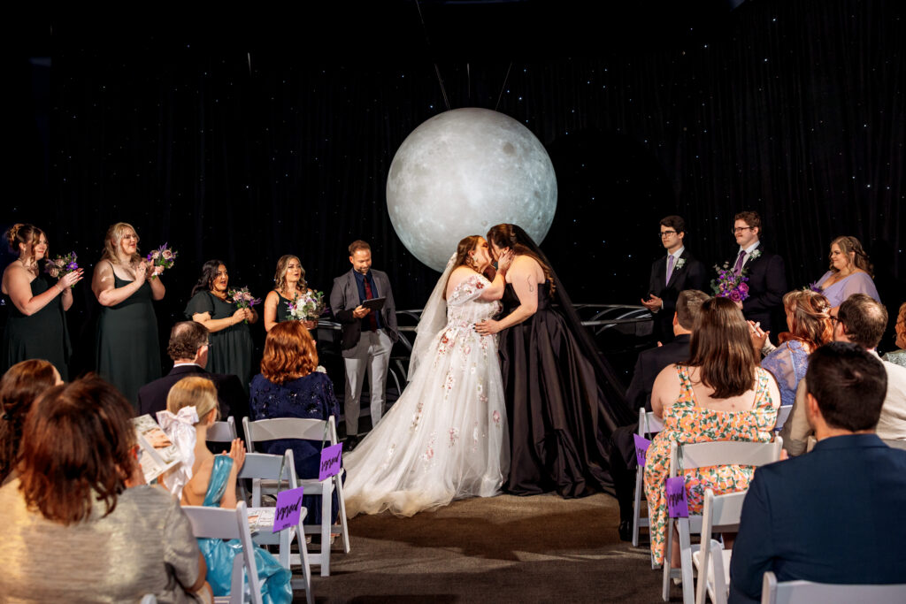 Two brides — one in a floral-embroidered white gown, the other in a black gown — share a kiss at the altar under a large illuminated moon backdrop against a starry black sky, as guests and a wedding party look on.