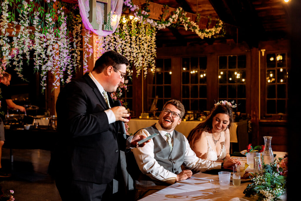 A groom speaks or reads at a table while a bride and another man laugh beside him, string lights and greenery decorating the rustic venue overhead.