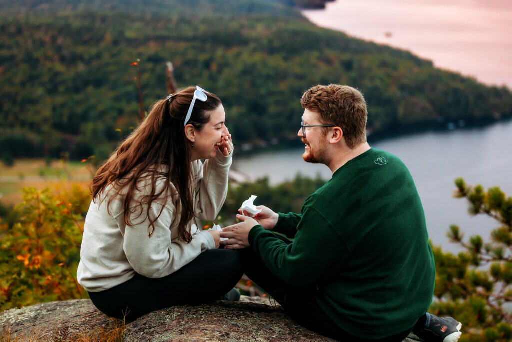 A couple sits close together on a rocky overlook at sunset, smiling at each other with a lake and forested hills in the background.