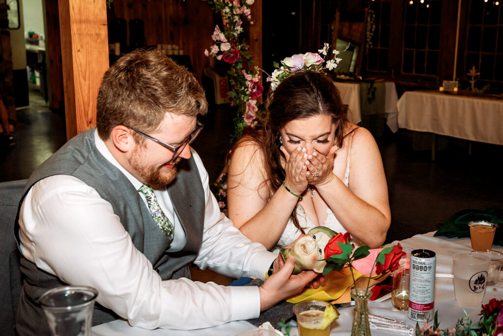 A bride reacts with an emotional, covered-mouth expression while the groom looks on smiling, both seated at a table set with drinks.