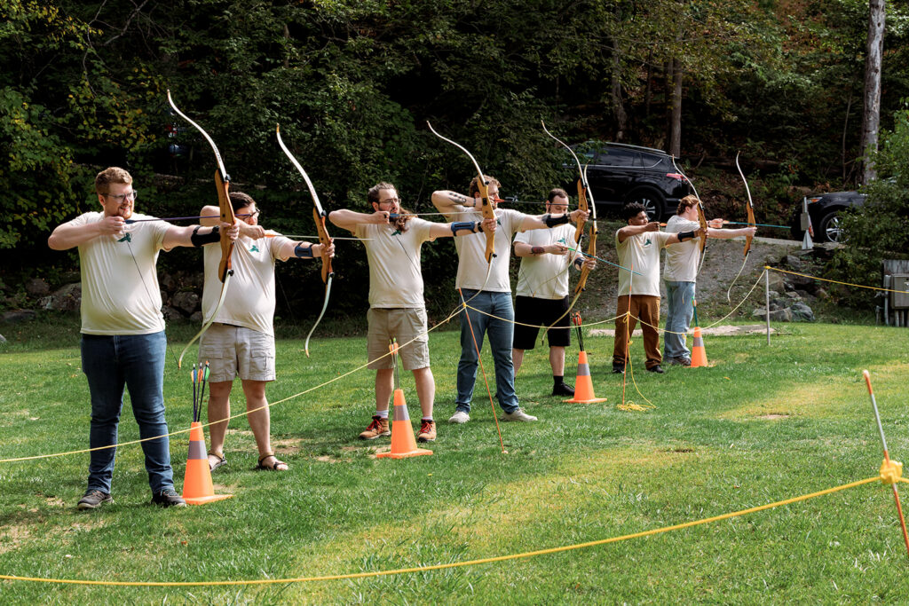 A line of people in matching t-shirts practice archery on a grassy lawn, aiming bows toward orange traffic cone targets.