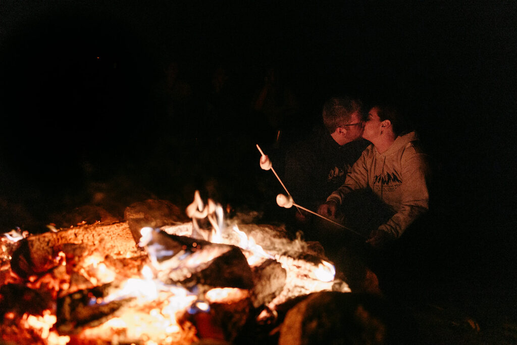 Two people roast marshmallows over a glowing campfire at night.