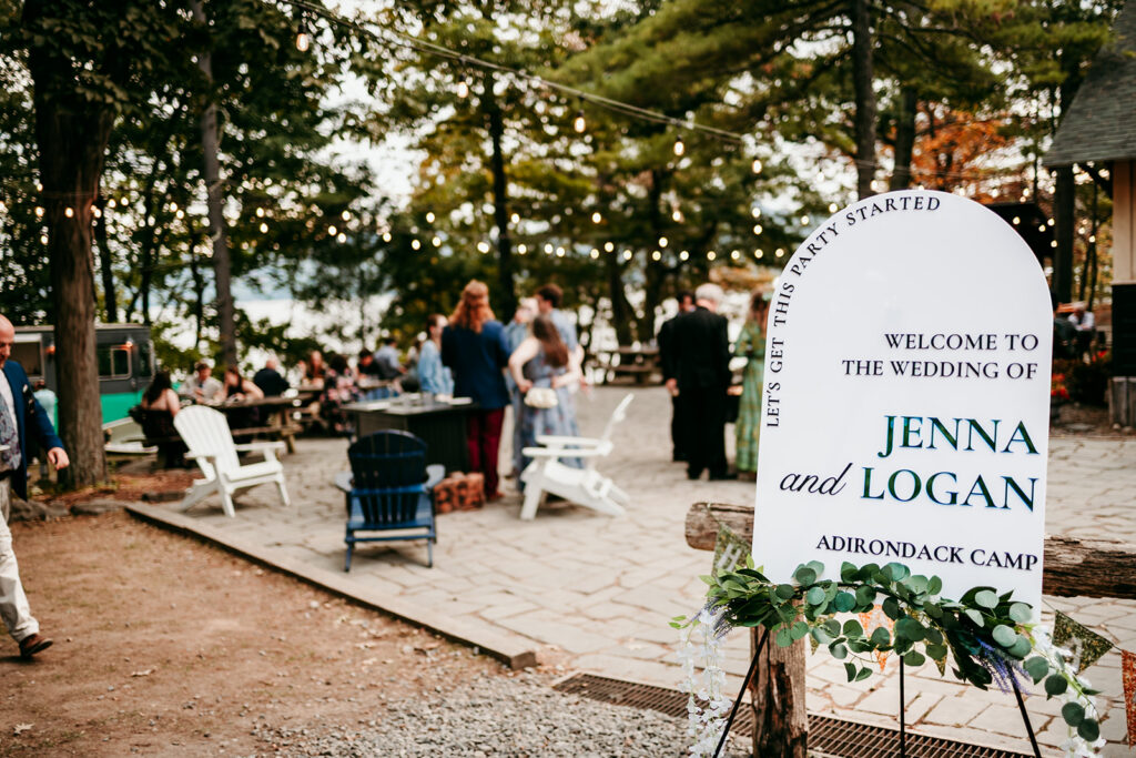 A wooden welcome sign reading "Welcome to the Wedding of Jenna and Logan, Adirondack Camp" stands outdoors near a lakeside gathering of guests.