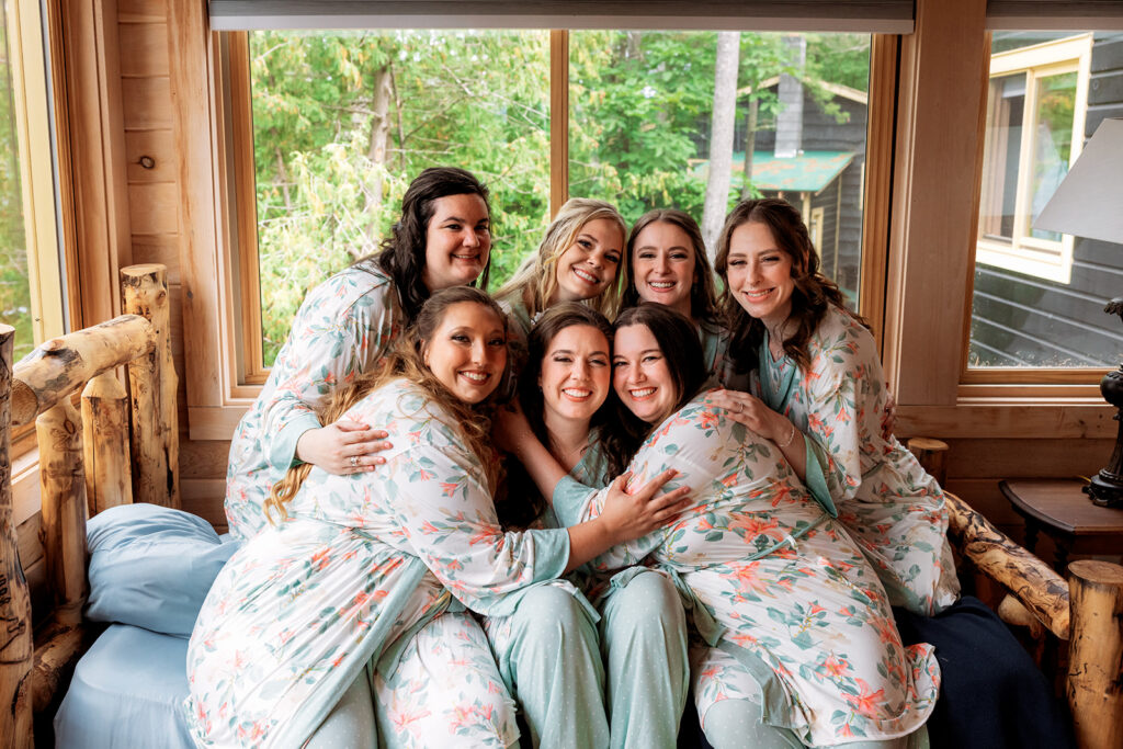 A group of women in matching floral pajama sets pose together laughing in a rustic cabin bedroom.