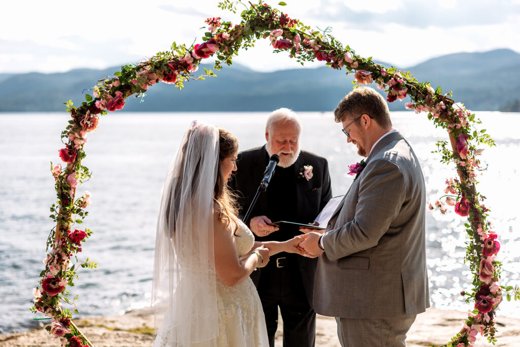 A bride and groom hold hands and exchange vows under a flower-covered arch with an officiant, ocean and mountains visible behind them.