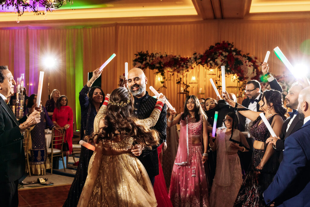Multi-day Indian wedding: the groom in ornate traditional attire is embraced and celebrated by dancing guests waving glow sticks under a floral-decorated stage.