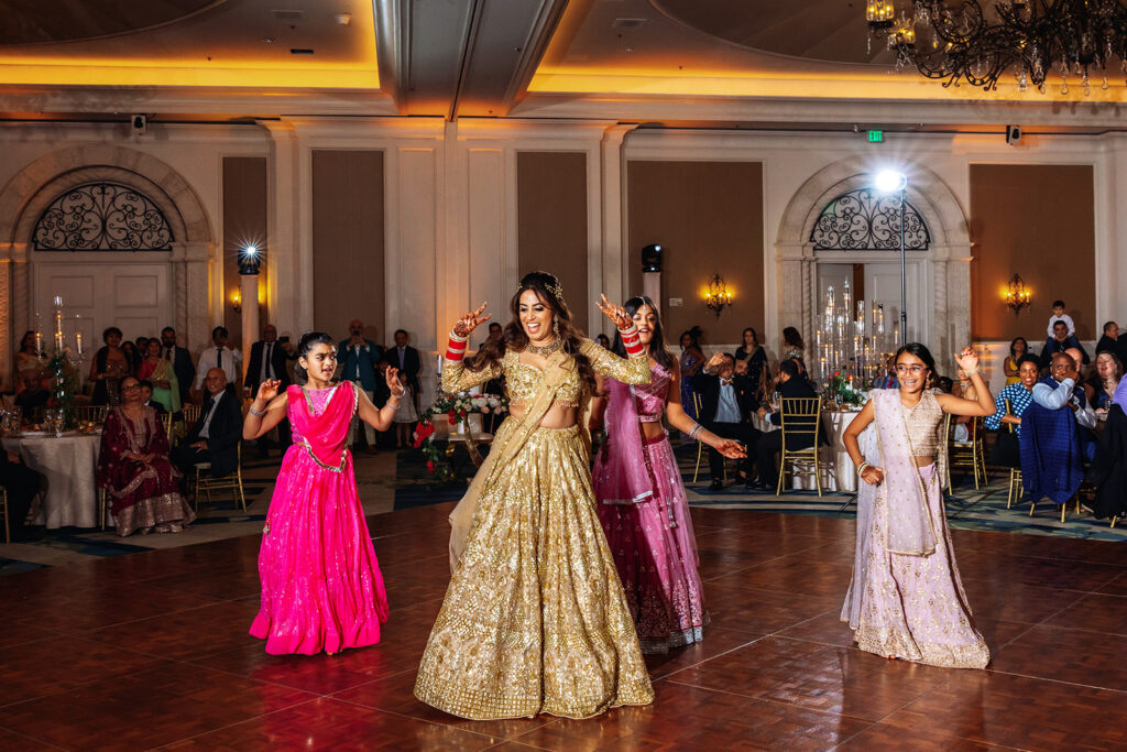 Multi-day Indian wedding: three women in vibrant pink, gold, and cream lehengas dance together on a dance floor as guests watch.