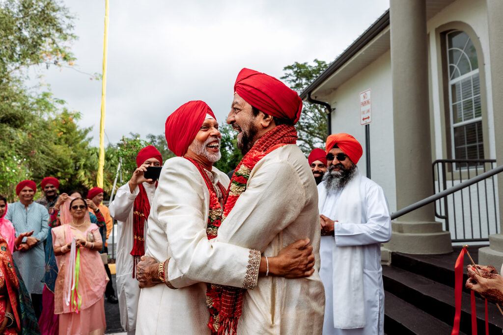 Multi-day Indian wedding: two men in red turbans and cream sherwanis embrace joyfully outside a venue, surrounded by family in traditional wedding attire.