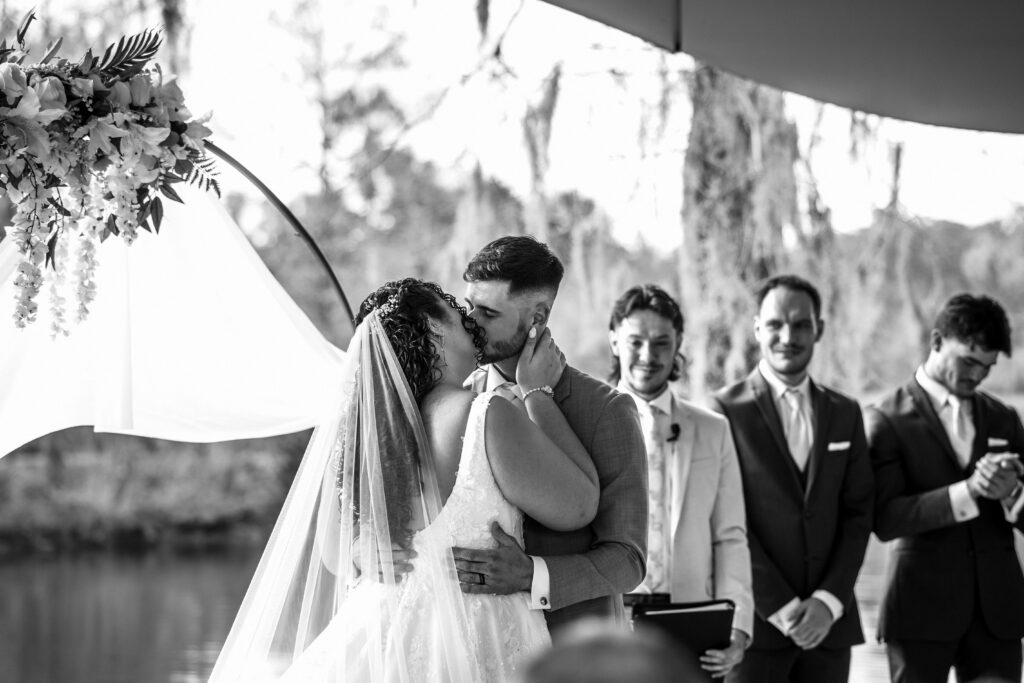 Black and white photo of the couple sharing a kiss at the altar, officiant and groomsmen looking on, with Spanish moss draped trees in the background.