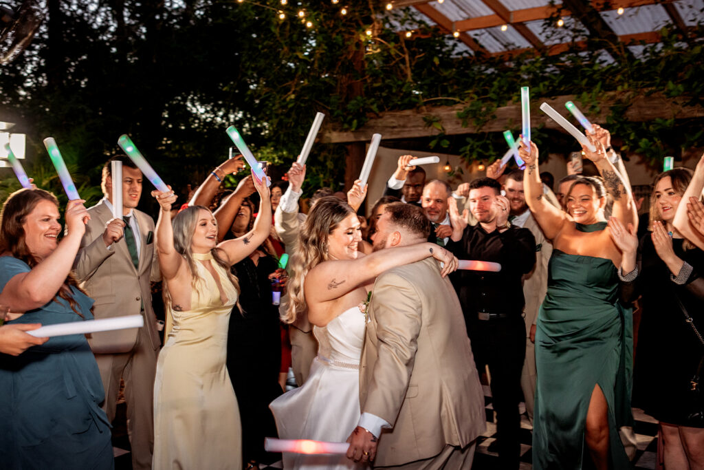 A bride in a strapless white gown and groom in a tan suit embrace and kiss on a checkered dance floor at their evening wedding reception, surrounded by cheering guests waving colorful glow sticks in the air.