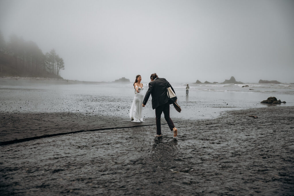 Groom stepping barefoot across tide pool toward bride during Ruby Beach Elopement, Olympic National Park, Washington