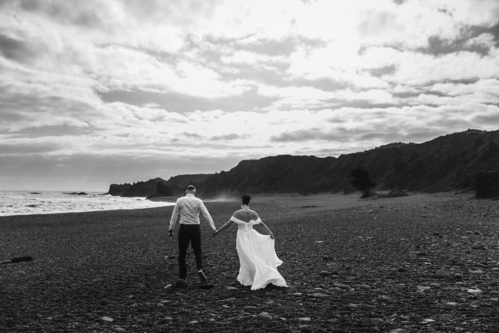 A couple walks hand in hand along a black sand beach during their Iceland elopement, with dramatic cliffs and crashing waves in the background.