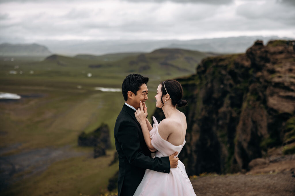 A couple shares an intimate moment on a cliffside overlook during their Iceland elopement, with sweeping green highlands stretching behind them.