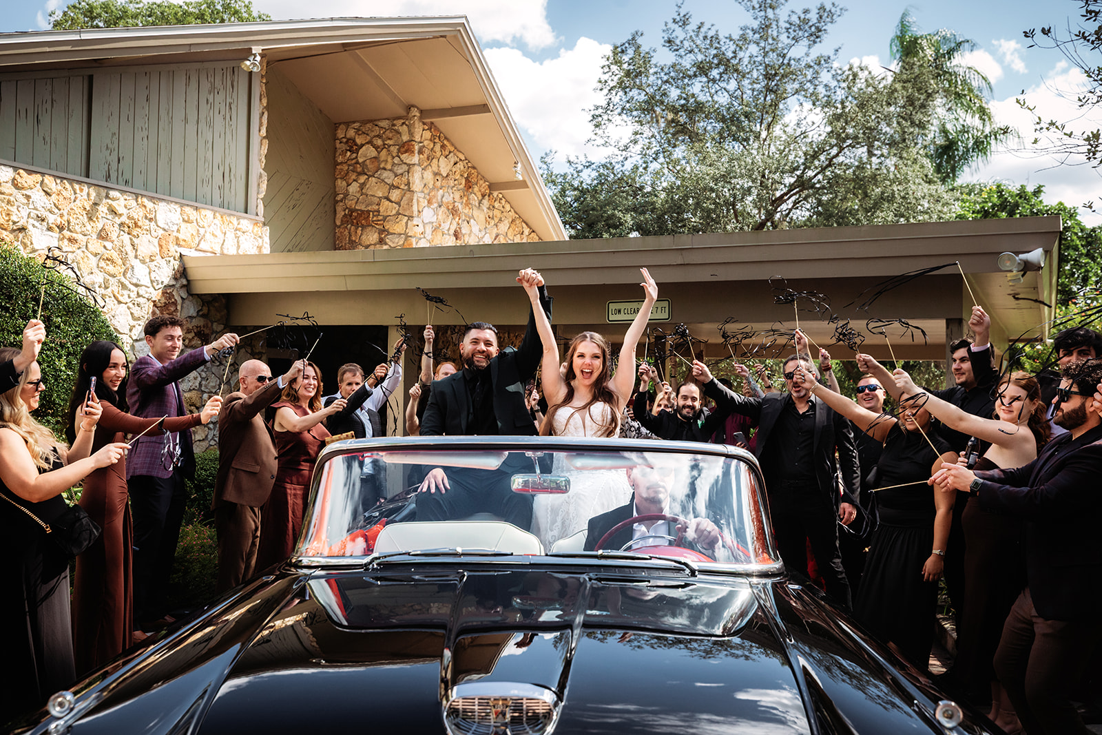 Newlyweds cheering with raised arms in a black vintage convertible as guests wave ribbon wands during their exit at Azalea Lodge at Mead Botanical Garden.